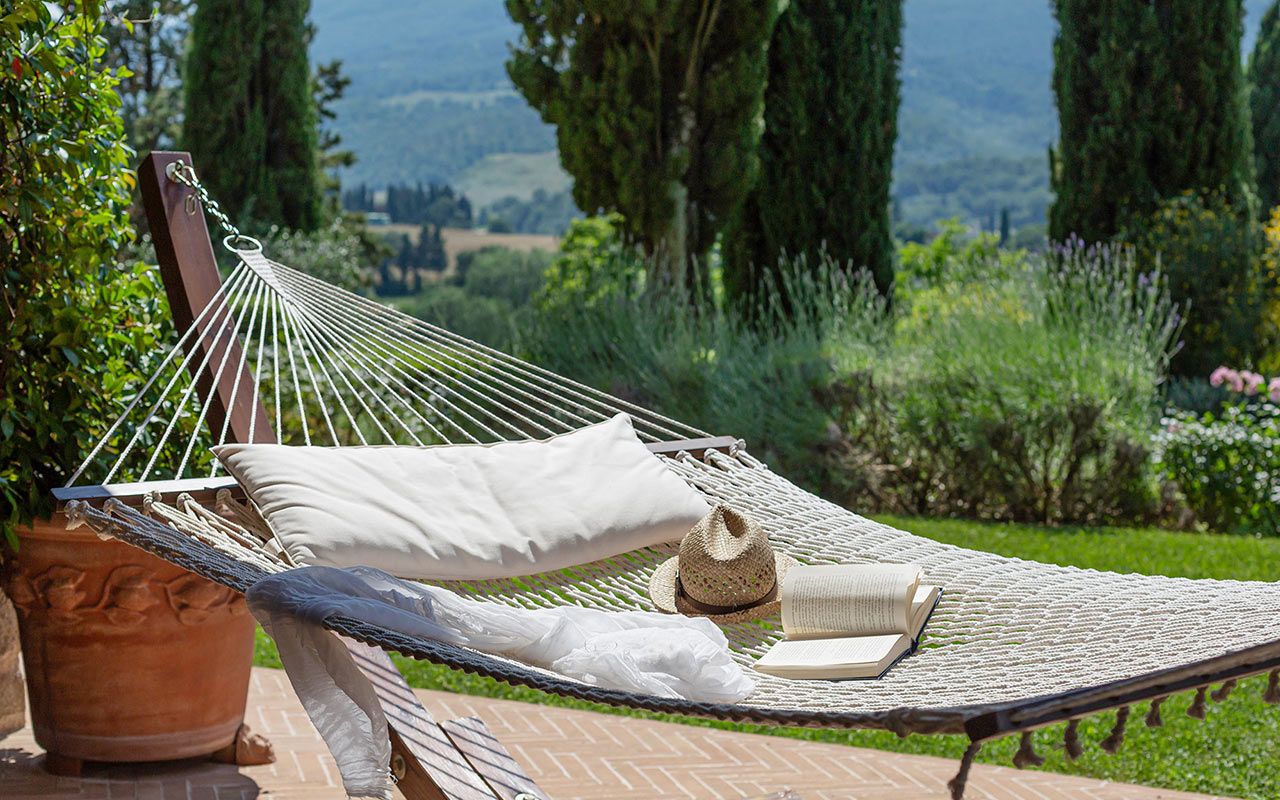 Amaca con cuscino, cappello e libro affacciata su un giardino rigoglioso e colline panoramiche.