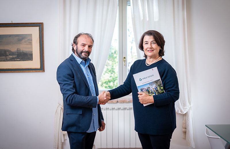 Due persone si stringono la mano in ufficio, tenendo una brochure Great Estate.