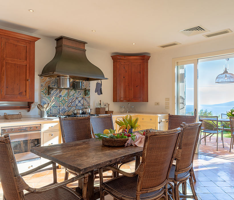 Cucina rustica con mobili in legno, piastrelle decorate e vista mare da porte a vetri.