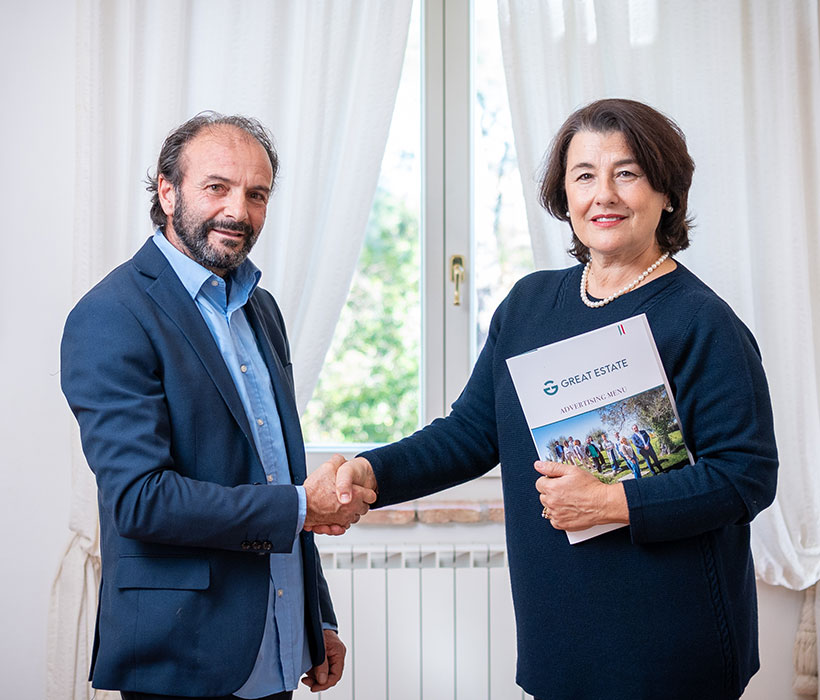 Due professionisti si stringono la mano vicino a una finestra, donna con brochure Great Estate.