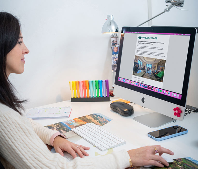 Donna lavora a una scrivania ordinata con iMac, penne colorate e sito Great Estate sullo schermo.