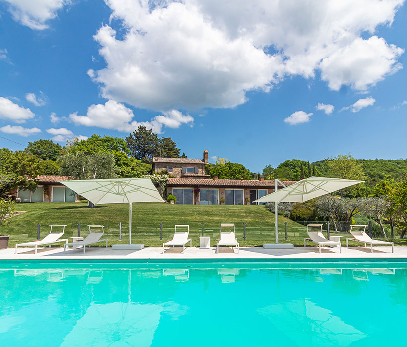 Villa di lusso con piscina, lettini e ombrelloni in un paesaggio verde e tranquillo.